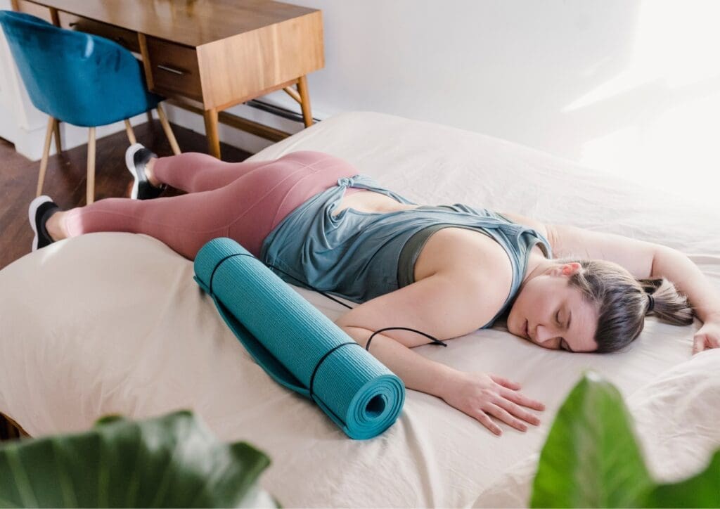Vrouw ligt met ogen dicht (voorover gevallen) op bed, met een yogamatje naast zich