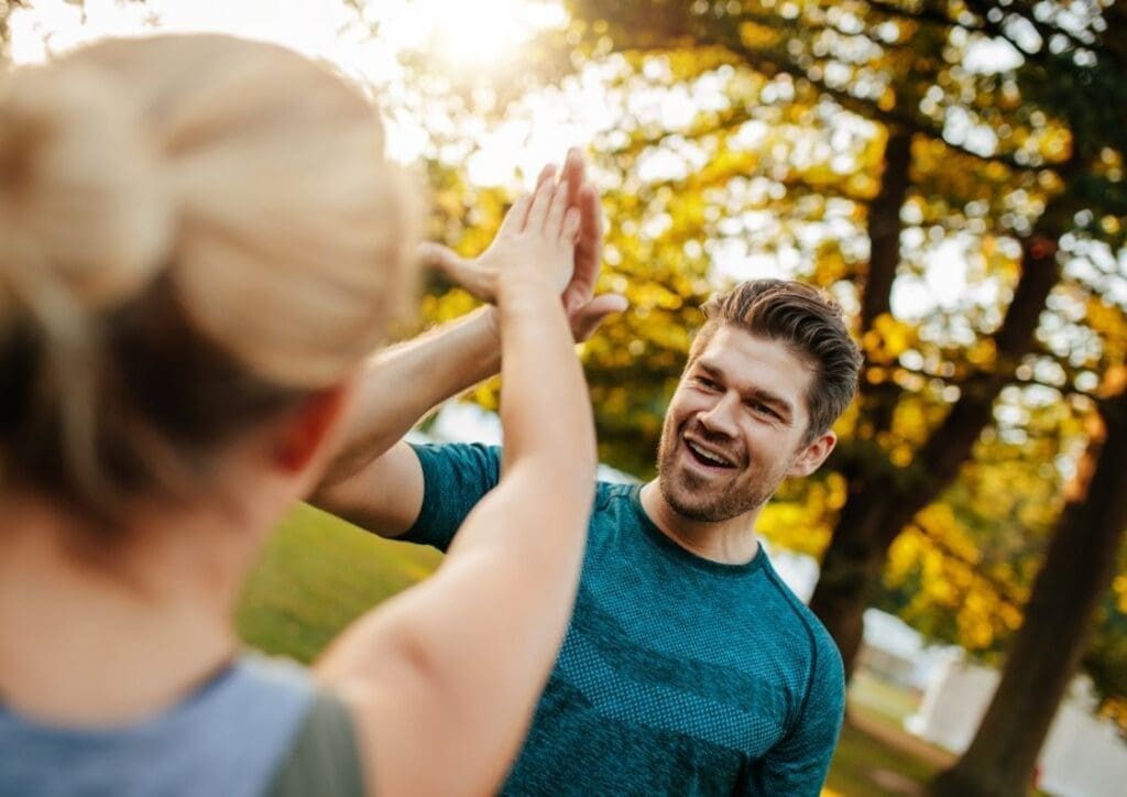 Lachende man en vrouw in sportkleding geven elkaar buiten een high five. Vrouw staat met haar rug naar camera. Op de achtergrond staan bomen. De zon schijnt er doorheen.